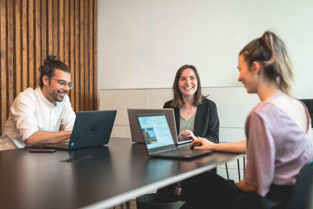 Drei Personen sitzen mit Laptops an einem Tisch und arbeiten gemeinsam, symbolisiert moderne Zusammenarbeit im Sinne von New Work